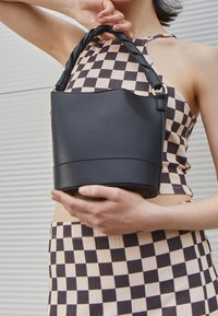 Osoba ubrana w czarno-beżowy kraciasty top i spódnicę, trzymająca czarną skórzaną torebkę z skręcanym uchwytem na tle ściany w paski.