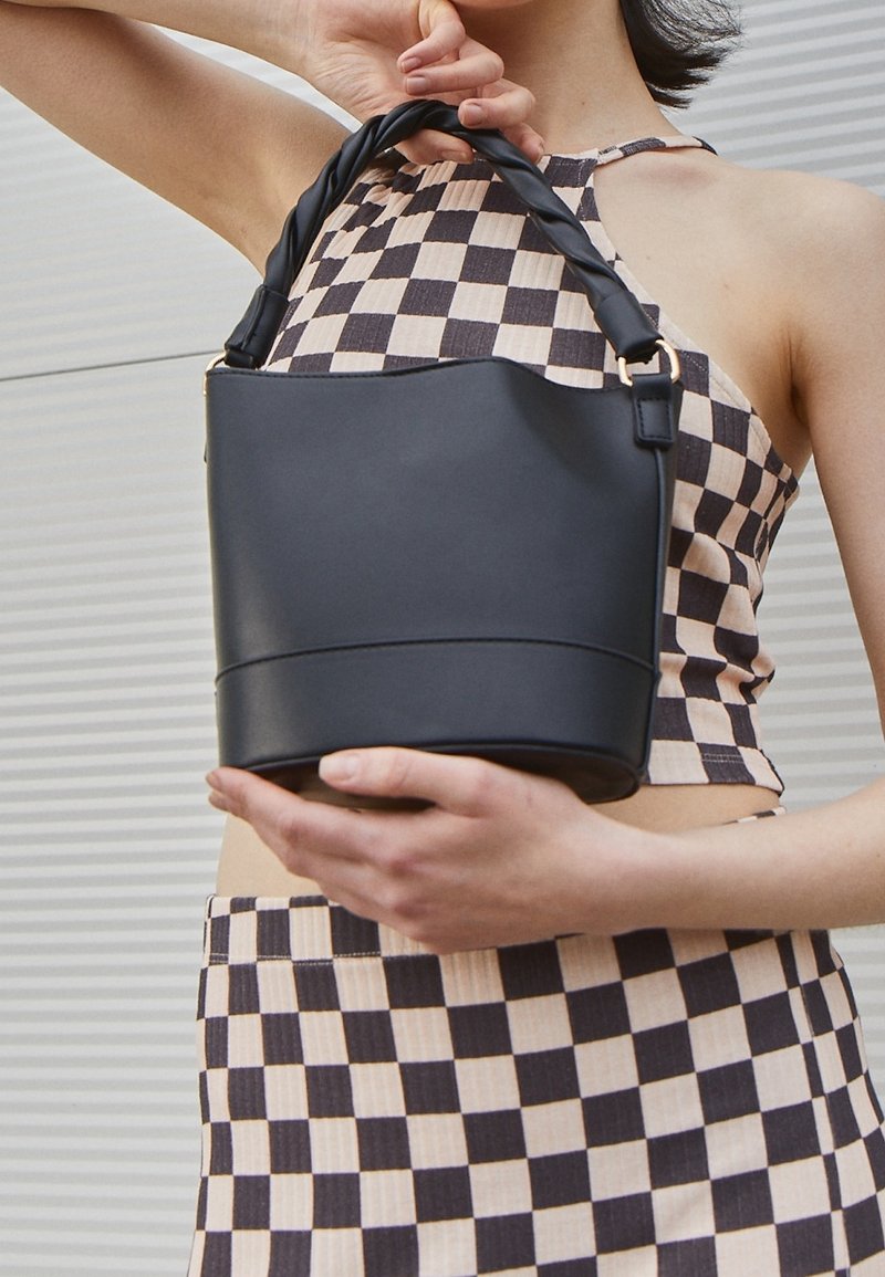 Osoba ubrana w czarno-beżowy kraciasty top i spódnicę, trzymająca czarną skórzaną torebkę z skręcanym uchwytem na tle ściany w paski.