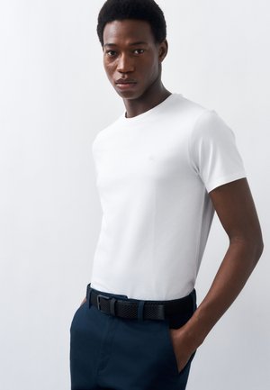 Ung mand står med hænderne i lommerne iført en ensfarvet hvid kortærmet t-shirt og mørkeblå bukser mod en hvid baggrund.