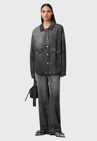 Donna che indossa una giacca di jeans nera oversize e dei pantaloni larghi abbinati, mentre tiene in mano sinistra una borsa a cintura nera.