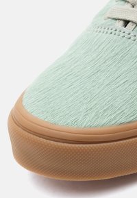 Gros plan sur l'avant d'une basket vert clair avec un tissu texturé et une semelle en caoutchouc beige, montrant les détails cousus et les lacets.