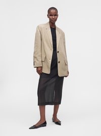 Γυναίκα που φοράει μπεζ oversized σακάκι, μαύρο τοπ, διάφανη μαύρη μίντι φούστα και μαύρες μυτερές μπαλαρίνες, στέκεται μπροστά σε απλό φόντο.