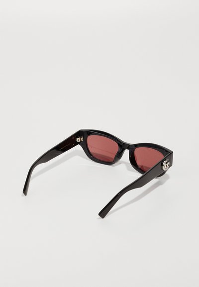 Gafas de sol de plástico negro con montura angular, lentes tintados de rojo y un detalle de logo plateado en el lateral. Textura suave, diseño moderno.