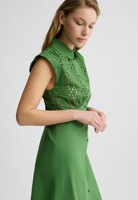 Abito verde con colletto, maniche corte e intricati motivi ritagliati nella parte superiore, mentre la parte inferiore è liscia e di un colore uniforme.