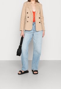 Blazer beige de doble botonadura, top de satén rojo, jeans de cintura alta de color azul claro, sandalias planas negras, sosteniendo un pequeño bolso negro de hombro.