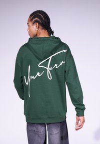 Haut à capuche vert avec une poche kangourou, arborant un grand texte en écriture cursive blanche "À ton tour" au dos. Tissu doux et coupe décontractée.