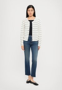 Witte cardigan met horizontale zwarte en gekleurde strepen, ronde halslijn en knopen, gecombineerd met donkerblauwe wijde jeans en zwarte ballerina's.