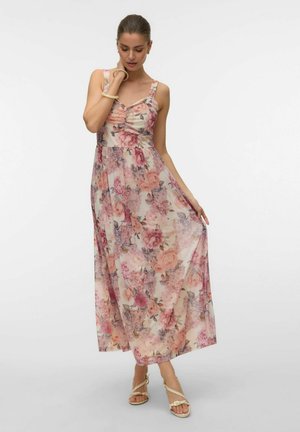 Kvinne iført en ermeløs, rosa blomstermønstret maxikjole og beige sandaler, som forsiktig holder i kjolens kant mens hun ser ned.