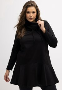Tunica nera con scollo a cerniera, maniche lunghe, orlo svasato e tasche frontali; realizzata in morbido cotone testurizzato.
