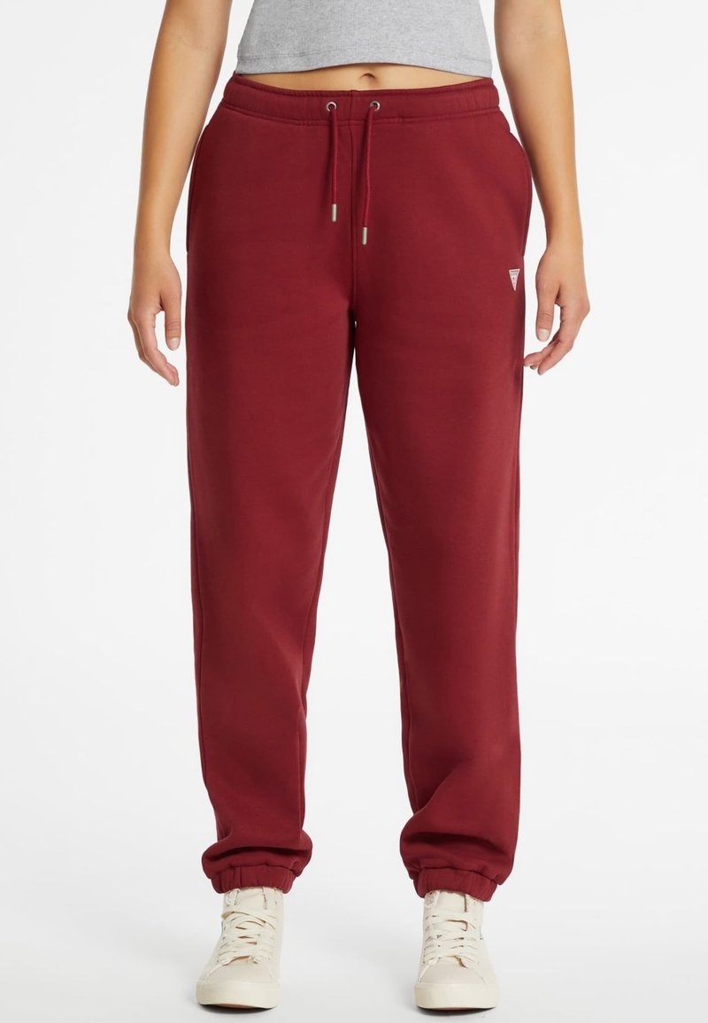 Mujer vestida con pantalones de chándal color burdeos con puños elásticos y zapatillas blancas, de pie contra un fondo blanco liso.