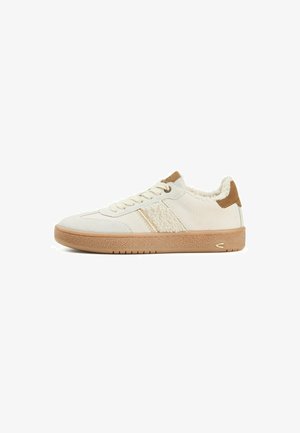 Lage beige sneaker met gumrubberen zool, vetersluiting aan de voorkant, schapenfleece voering en een bruine suède hielaccent, zijaanzicht op een witte achtergrond.