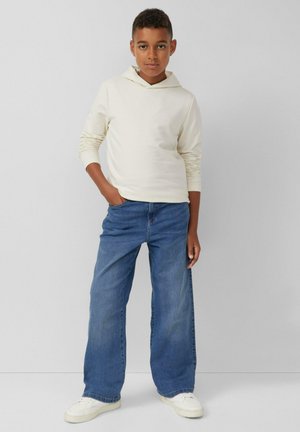 Felpa a maniche lunghe color crema con cappuccio regolabile, abbinata a jeans a gamba larga blu chiaro e sneakers bianche, su uno sfondo neutro.