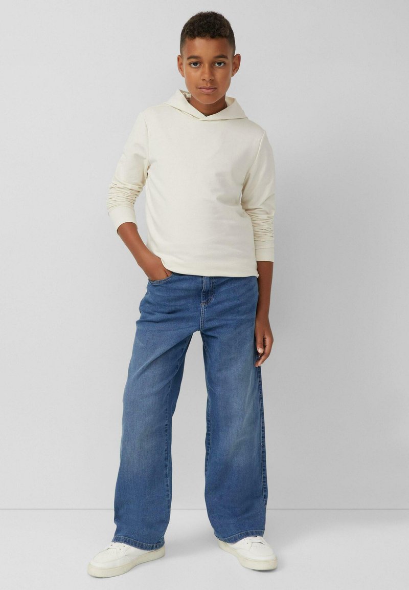 Felpa a maniche lunghe color crema con cappuccio regolabile, abbinata a jeans a gamba larga blu chiaro e sneakers bianche, su uno sfondo neutro.