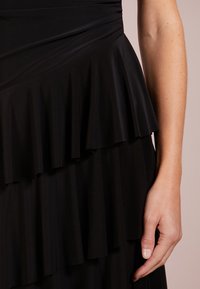 Svart klänning med lager av volanger, slät textur och en figurnära bodice. Tyget är lätt och faller mjukt.