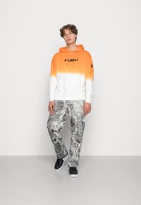 Hætte sweatshirt i gradient orange og hvid med sort "FUBU" logo, parret med grå marmor-mønstrede bukser og sorte sneakers.