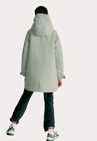 Parka lunga verde chiaro con cappuccio, polsini elasticizzati e taglio dritto, abbinata a jeans scuri e sneakers con dettagli verdi.