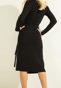 Robe en maille côtelée noire avec des manches longues, une taille ceinturée et une fente sur le côté, mettant en valeur une silhouette ajustée et une texture lisse.