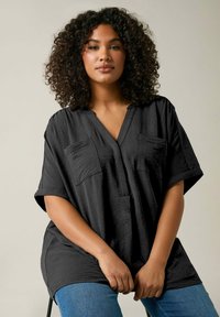 Tunic nera a maniche corte con vestibilità ampia, dotata di due tasche sul petto e scollatura a V. Realizzata in tessuto testurizzato.