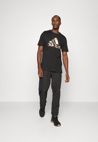 Camiseta de algodón negra con un logo de Adidas en dorado y floral, combinada con pantalones negros con rayas azules y zapatos negros.