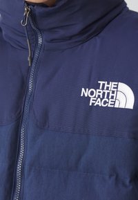 Veste bleu marine avec fermeture éclair et col montant, avec le logo brodé blanc "THE NORTH FACE" sur la poitrine.