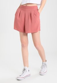 Růžové plisované šortky s širokým pásem, vyrobené z hladké tkaniny. Stylizované s bílými vysokými teniskami. Bez viditelných vzorů nebo kovových doplňků.