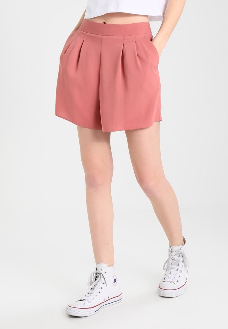 Růžové plisované šortky s širokým pásem, vyrobené z hladké tkaniny. Stylizované s bílými vysokými teniskami. Bez viditelných vzorů nebo kovových doplňků.