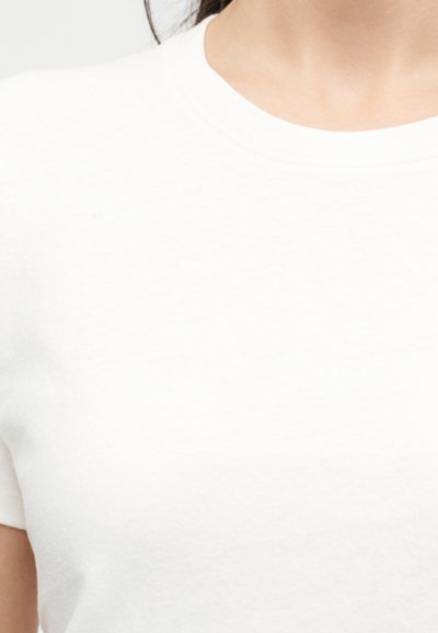 Κοντινό πλάνο ενός ατόμου που φοράει ένα απλό λευκό T-shirt με στρογγυλή λαιμόκοψη, δείχνοντας το επάνω μέρος του στήθους, τον λαιμό και μέρος του αριστερού βραχίονα.