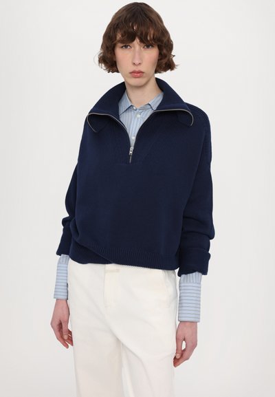 Navy knitted sweater with a high collar and zip closure, layered over a light blue striped shirt, paired with white trousers.