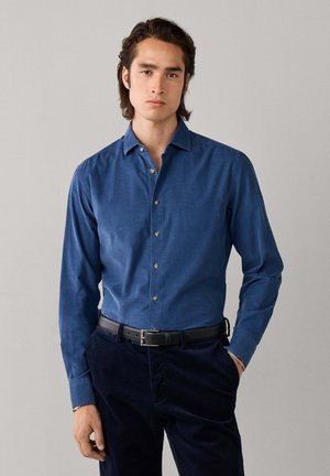Man met halflang donker haar die een blauw overhemdbout draagt, ingestopt in een donkere broek met een zwarte riem, staat tegen een effen achtergrond.