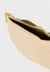 Béžová kožená kabelka do ruky s zaobleným tvarem, zlatým zipem a zelenou podšívkou. Obsahuje odnímatelnou sponu pro univerzální nošení.