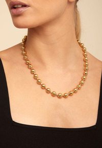 Collar de cuentas de oro que presenta cuentas esféricas de tamaño uniforme, con una textura pulida, llevado sobre un cuello desnudo contra una blusa negra.