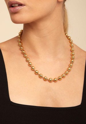 Collar de cuentas de oro que presenta cuentas esféricas de tamaño uniforme, con una textura pulida, llevado sobre un cuello desnudo contra una blusa negra.