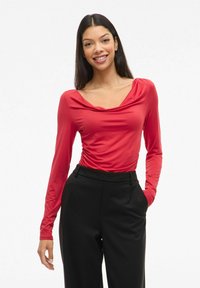 Czerwona bluzka z długim rękawem i dekoltem w łódkę, o gładkiej teksturze i detalach marszczenia po bokach, w połączeniu z czarnymi spodniami o wysokim stanie.