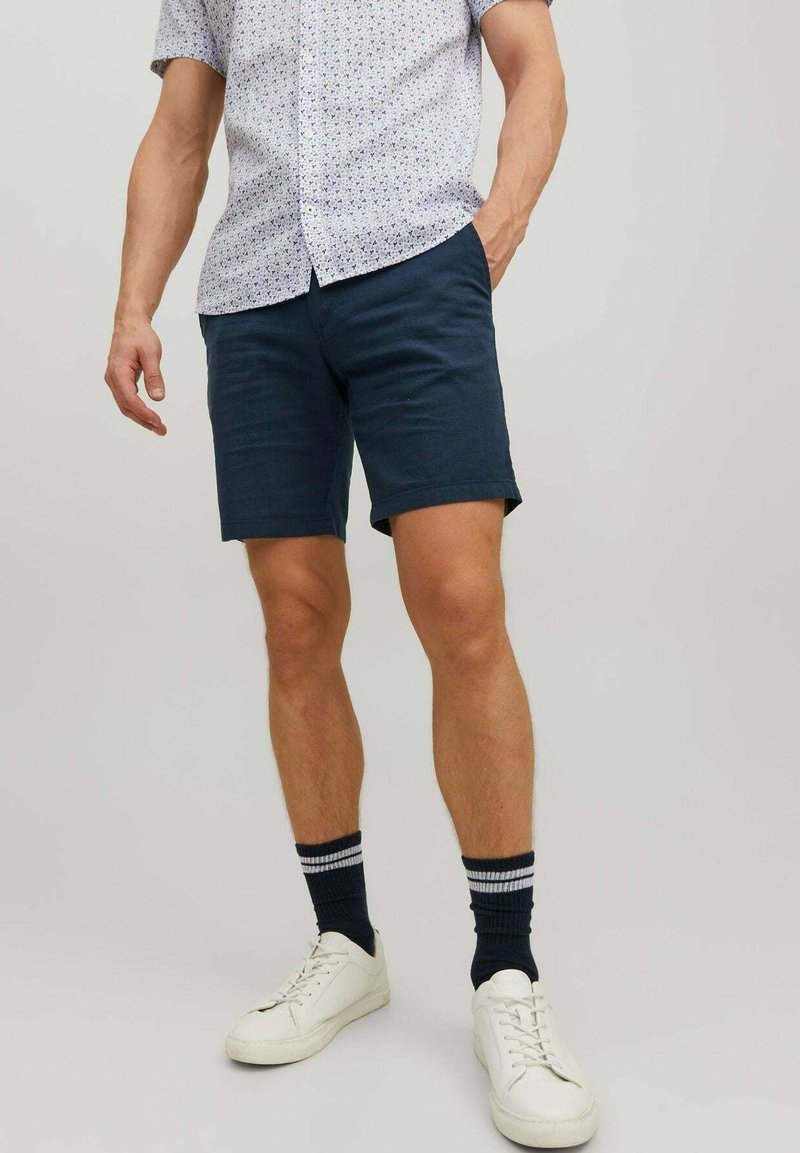 Shorts bleumarin din bumbac, cu o textură fină, asortați cu o cămașă cu mânecă scurtă, cu model, și adidași albi, purtând șosete în dungi închise.