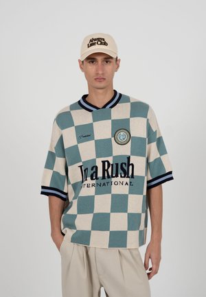 Fiatal férfi bézs és zöldeskék kockás ingben, amelyen az "In a Rush International" felirat olvasható, valamint egy bézs sapkában, amelyen az "Always Late Club" felirat található.