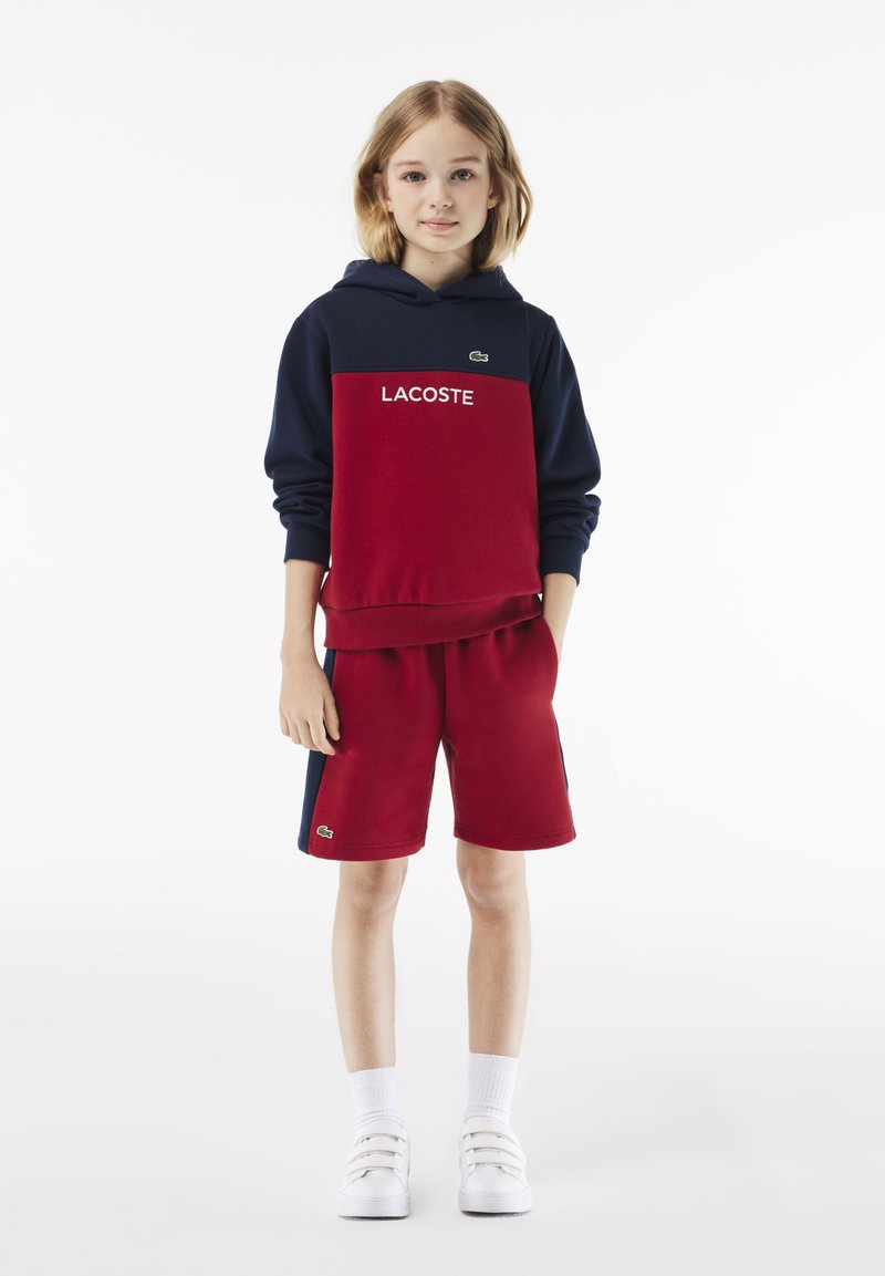 Zweifarbige Kapuzenjacke in Rot und Marineblau mit "LACOSTE"-Markenlogo, kombiniert mit passenden roten Shorts. Verfügt über gerippte Bündchen und seitliche Akzente, aus einem Baumwoll-Mix gefertigt.