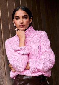 Rózsaszín vastag kötött pulóver magas nyakkal és visszahajtott mandzsettával. Barna fényes szoknya fölött viselve. Az anyag puhanak és texturáltnak tűnik.