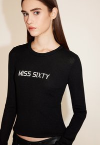 Schwarzes langärmliges Rippoberteil mit "MISS SIXTY" in weißem Text, runder Halsauschnitt und figurbetontem Design. Das Model trägt es mit schwarzen Hosen.