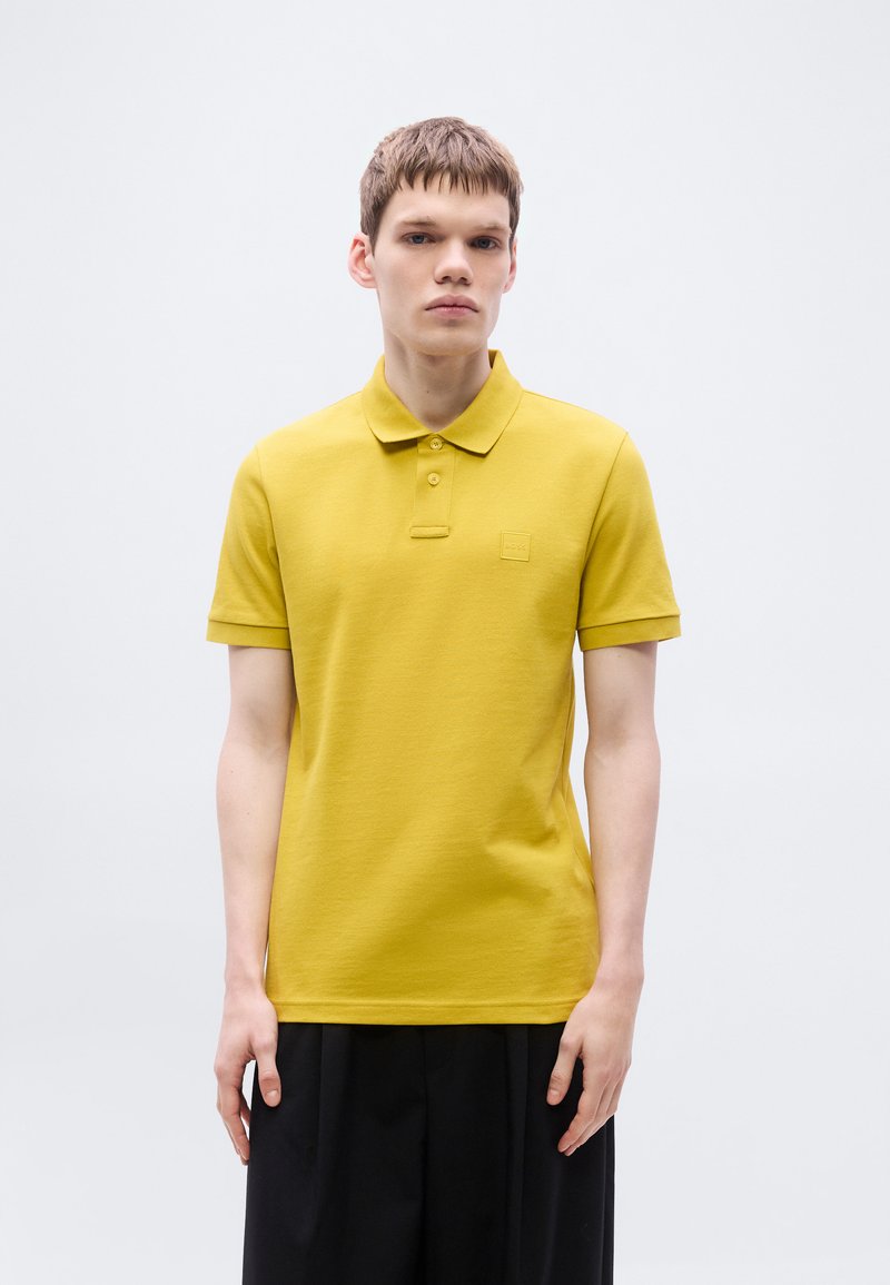 Polo jaune avec un tissu texturé, une patte à trois boutons et une petite écusson logo sur la poitrine. Manches courtes et coupe standard.