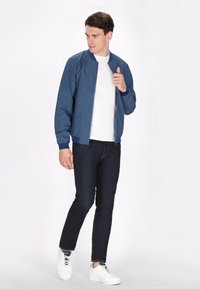 Giacca bomber navy con motivo geometrico, chiusura con zip, polsini e bordo a costine, abbinata a jeans scuri e sneaker bianche.