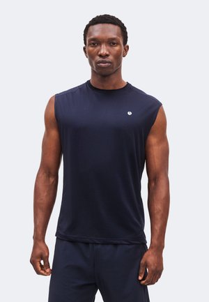 Homme musclé debout de face, portant un débardeur bleu marine et un short assorti sur un fond uni.