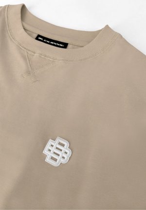 Beżowa bluza z okrągłym dekoltem, prążkowanym kołnierzem, przeszytym detalem w kształcie litery V i białym logo z splecionymi literami na przedniej części klatki piersiowej.