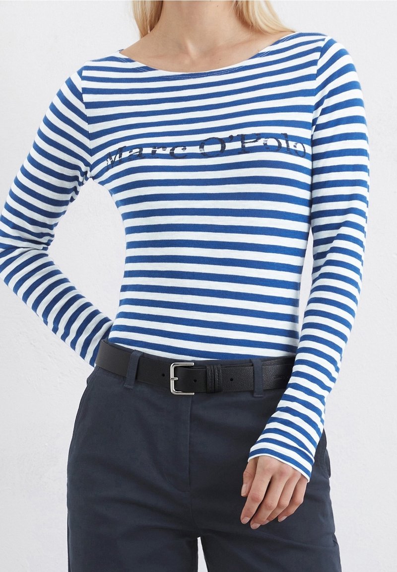 Niebieska i biała w paski koszula z długim rękawem, z dekoltem w łódkę, z logo nadrukowanym na piersi. Noszona z ciemnymi spodniami i czarnym paskiem.