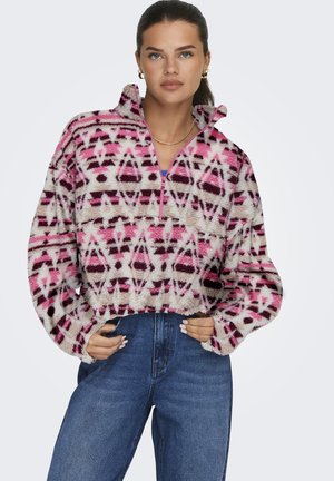 Junge Frau mit dunklem Haar, die goldene Creolen und eine Halskette trägt, einen pinkfarbenen, gemusterten Fleece-Pullover und hohe, blaue Jeans.