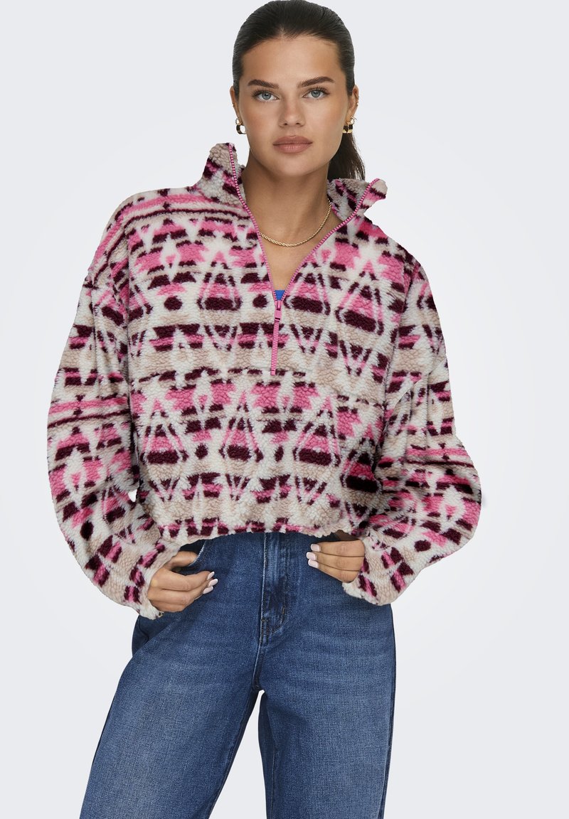 Junge Frau mit dunklem Haar, die goldene Creolen und eine Halskette trägt, einen pinkfarbenen, gemusterten Fleece-Pullover und hohe, blaue Jeans.