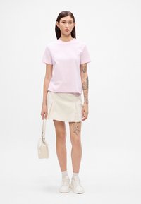 Lyserød bomulds T-shirt med korte ærmer, parret med en cremefarvet nederdel. Modellen bærer hvide sneakers og holder en lille cremefarvet taske. Synlige tatoveringer på armen.