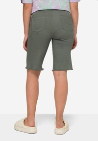 Bermudas de mezclilla verde con dobladillo deshilachado, dos bolsillos traseros y un ajuste ceñido. Llevadas con zapatos de slip-on acolchados de color claro.