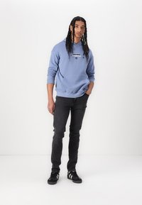 Light blue hoodie with a front pocket and graphic detail, paired with black jeans and black sneakers featuring white stripes.