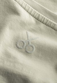 Lichtgrijze stof met een gladde textuur, met een geborduurde schaarontwerp in een lichtere tint. Close-up van de detail van het kledingstuk.