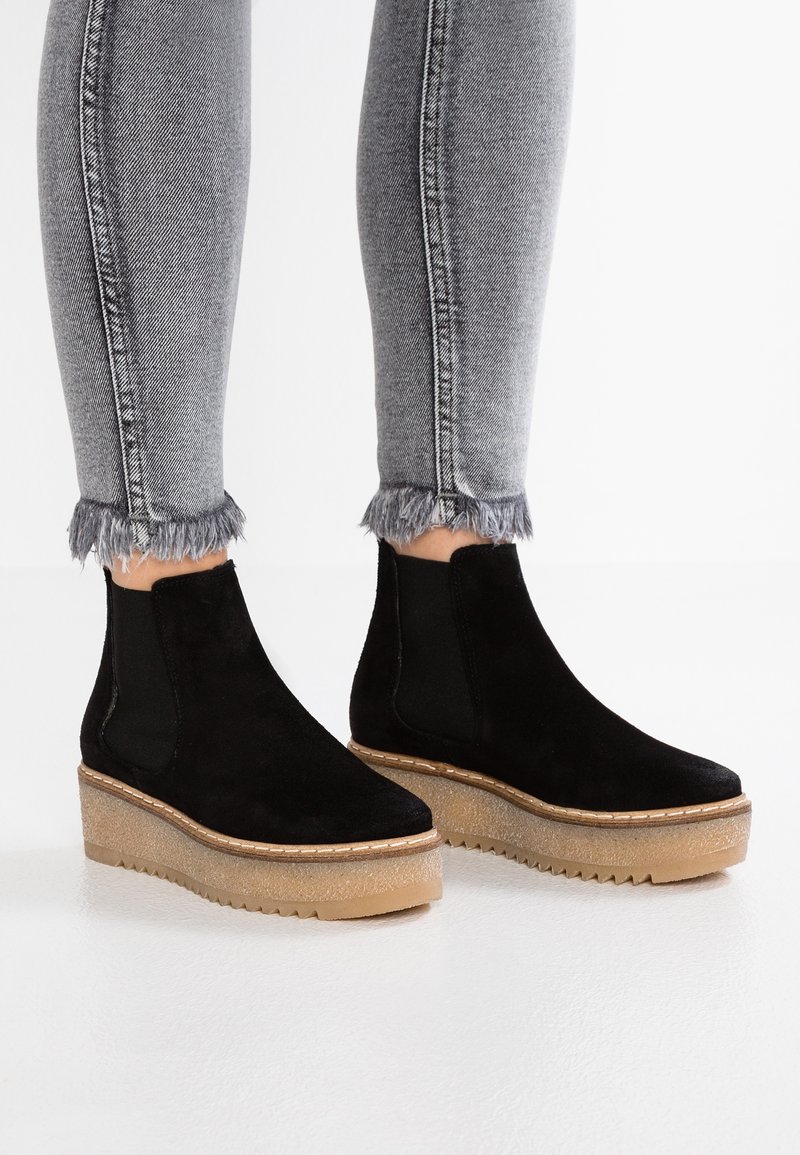 Jambes portant un jean gris à ourlet effiloché et des bottines compensées en daim noir avec semelles en caoutchouc couleur caramel sur fond blanc.